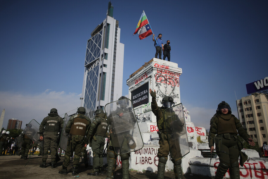 Dos personas protestan sobre la base del monumento al general Baquedano, mientras carabineros de Fuerzas Especiales rodean la estructura.
